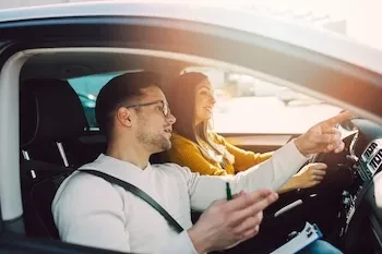 Junges Paar sitzt im Auto, der Mann zeigt mit dem Finger und spricht, während die Frau am Steuer lächelnd zuhört.