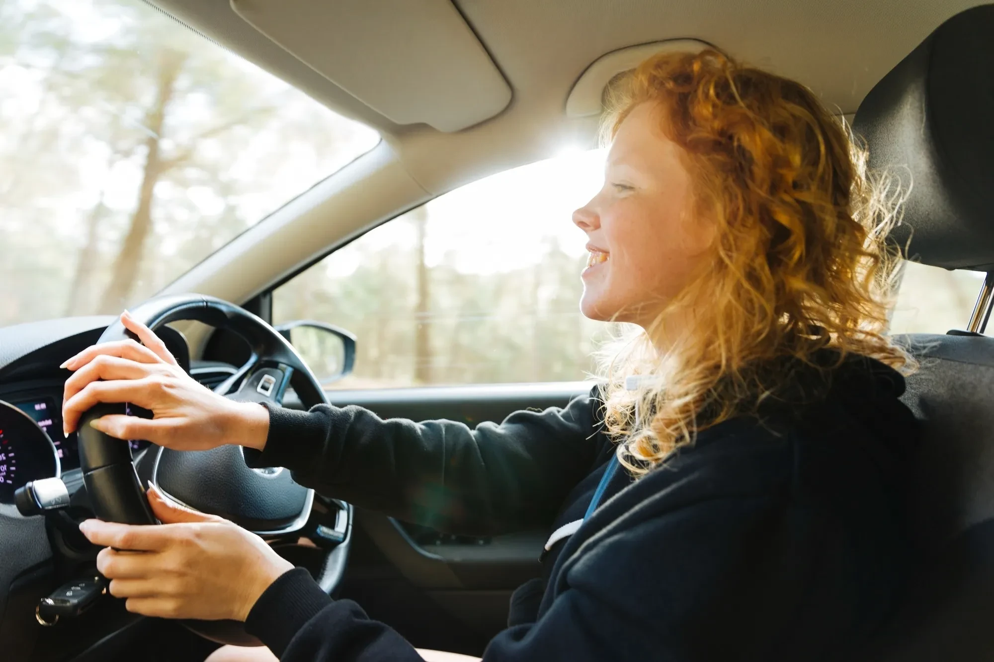 Junge Fahrschülerin mit lockigem Haar sitzt lächelnd am Lenkrad eines Autos.