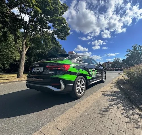 Schwarzes Audi-Fahrschulauto der Fahrschule Fahrtwind mit grünen Akzenten am Straßenrand unter blauem Himmel mit Wolken.