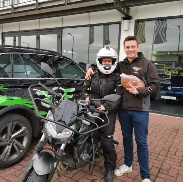Zwei Männer posieren vor einem Motorrad und einem schwarzen Auto, einer trägt Motorradhelm und Ganzkörperkleidung, der andere hält ein Brot und trägt eine schwarze Jacke.