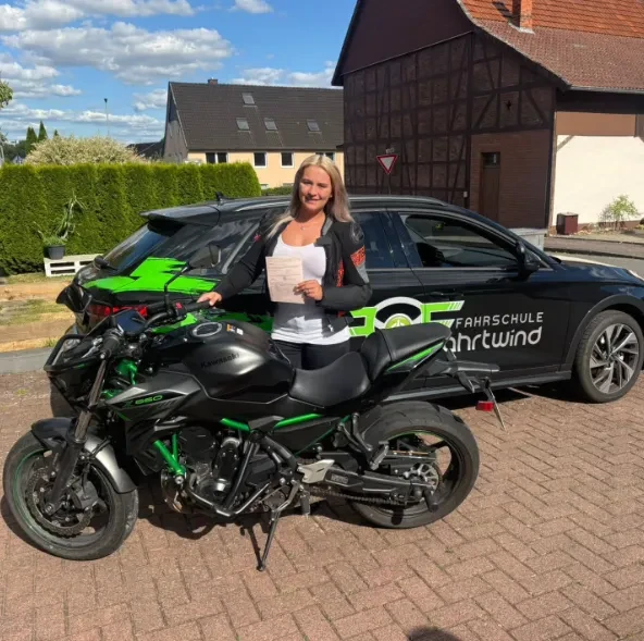 Frau steht mit einem Zertifikat neben einem grünen und schwarzen Motorrad, hinter ihr steht ein schwarzes Auto mit der Aufschrift Fahrschule Fahrtwind.